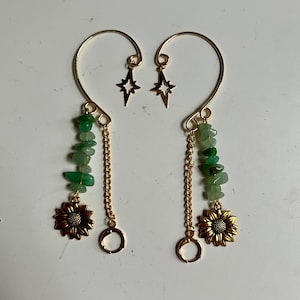 Puede incluir: Un par de puños de oreja dorados. Cada puño presenta una cadena colgante con virutas de piedras preciosas verdes, un dije de girasol y un pequeño anillo dorado. Arriba, se adjunta un dije de estrella dorada.