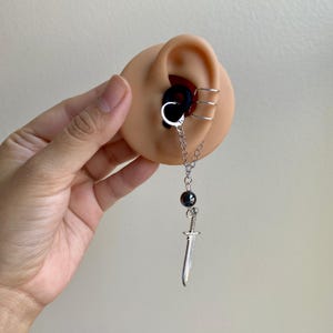 May include: A silver chain earring with a small sword pendant, a dark bead, and a silver hoop earring. The earring is displayed on a tan ear model with three silver ear cuffs.