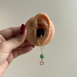 May include: A display of ear jewelry featuring a black plug, gold hoops, and a dangling earring. The dangling earring has a green bead and a gold star charm. The ear is a light peach color.