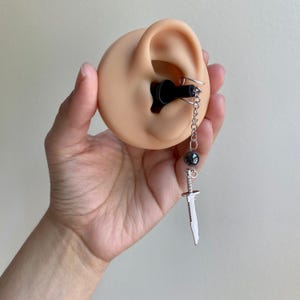 May include: A silver dagger earring with a black bead and chain detail. The earring is attached to a black ear plug, displayed on a prosthetic ear. The dagger has a detailed blade and handle.
