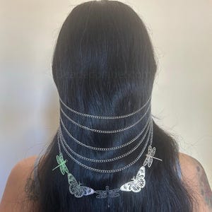 May include: Silver-toned head chain with multiple strands cascading down the back. The chain features dragonfly and butterfly charms. The dragonfly charms are light green. The head chain is worn over black hair.