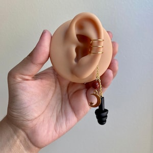 May include: Gold ear cuff with three rings and a dangling charm. The charm features a crescent moon and star, with red beads and a black earplug. The ear cuff is displayed on a prosthetic ear.