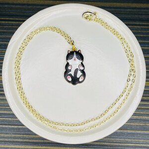 May include: A gold chain necklace with a black and white cat charm. The cat is curled up with its paws tucked in and has pink ears and nose.