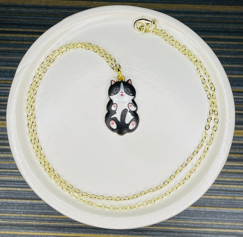 May include: A gold chain necklace with a black and white cat charm. The cat is curled up with its paws tucked in and has pink ears and nose.