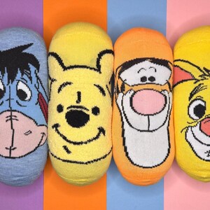 May include: Six colorful socks featuring cartoon characters. The socks display the faces of Owl, Eeyore, Winnie the Pooh, Tigger, Rabbit, and Piglet. The socks are arranged on a striped background of yellow, red, purple, orange, and green.