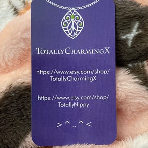 May include: Purple business card with white text and a white chain with a green gem pendant. The text reads "TotallyCharmingX" and includes two website addresses: "https://www.etsy.com/shop/TotallyCharmingX" and "https://www.etsy.com/shop/TotallyNippy".