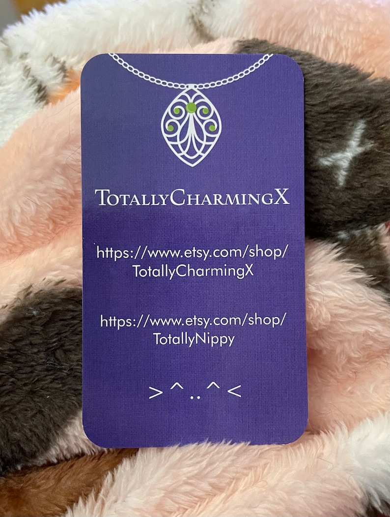 May include: Purple business card with white text and a white chain with a green gem pendant. The text reads "TotallyCharmingX" and includes two website addresses: "https://www.etsy.com/shop/TotallyCharmingX" and "https://www.etsy.com/shop/TotallyNippy".