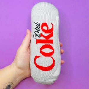 May include: A gray sock with the words "Diet Coke" in red script. The sock is designed to resemble a Diet Coke can. The background is a solid purple.