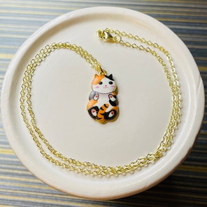 May include: A gold chain necklace with a charm of a calico cat with orange, black, and white fur. The cat is sitting with its paws tucked in and has a pink nose and white whiskers.