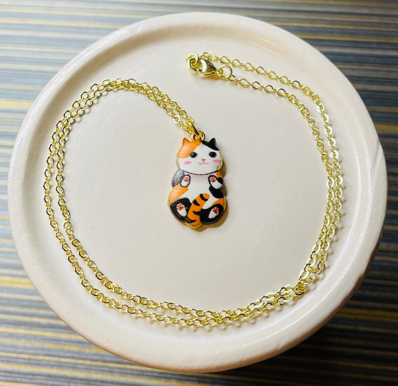 May include: A gold chain necklace with a charm of a calico cat with orange, black, and white fur. The cat is sitting with its paws tucked in and has a pink nose and white whiskers.