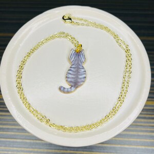 May include: A gold chain necklace with a gray and white cat pendant. The cat is facing away from the viewer and has a striped pattern.
