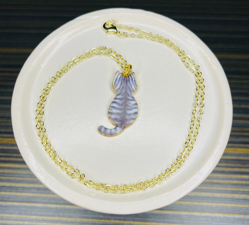 May include: A gold chain necklace with a gray and white cat pendant. The cat is facing away from the viewer and has a striped pattern.