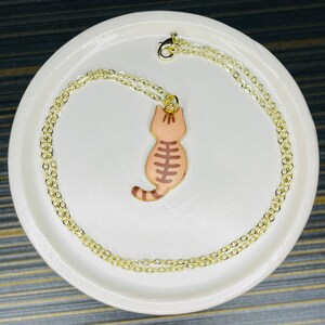May include: A gold chain necklace with a brown and white cat pendant. The cat is facing away from the viewer and has a striped tail.