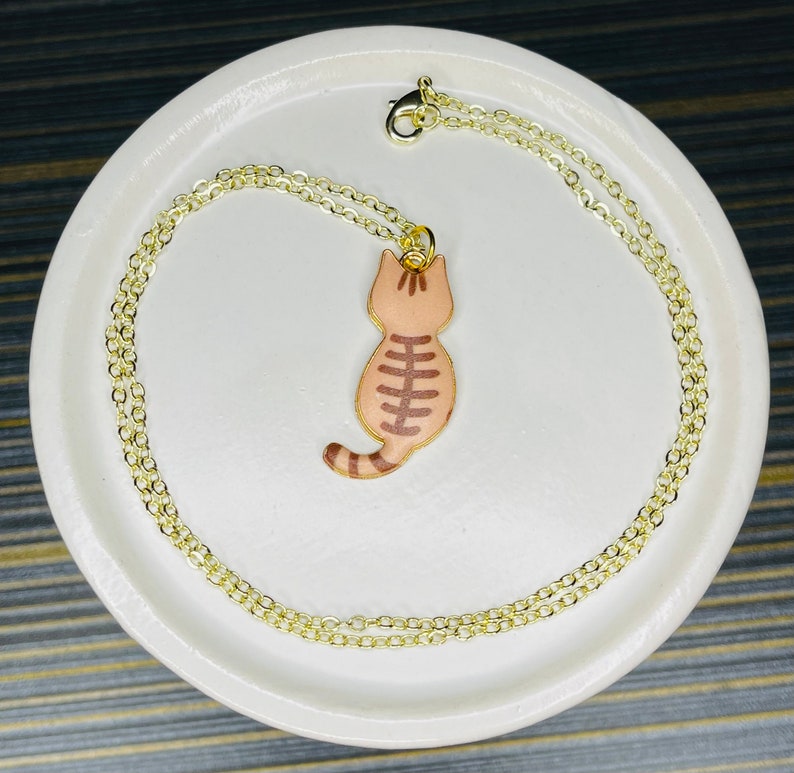 May include: A gold chain necklace with a brown and white cat pendant. The cat is facing away from the viewer and has a striped tail.