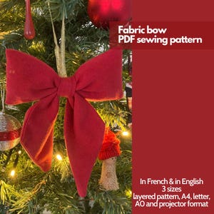 Peut inclure: Un nœud en tissu rouge suspendu à un sapin de Noël. Le nœud est fait d'un tissu doux et moelleux et a un design classique et élégant. L'image comprend le texte "Fabric bow PDF sewing pattern" et "In French & in English 3 sizes layered pattern, A4, letter, AO and projector format".