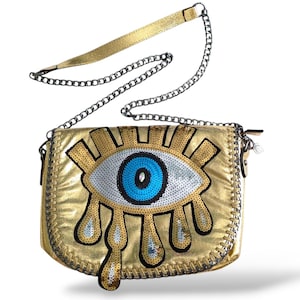 May include: Gold crossbody bag with an eye design. The bag has a metallic gold finish and a large eye embellished with blue, white, and silver sequins. It has a chain strap and a gold shoulder strap.