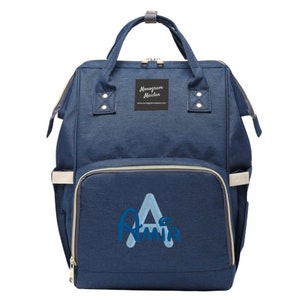 Op de afbeelding: Een marineblauwe luiertas met een voorvak en een handvat aan de bovenkant. De tas heeft de tekst "Monogram Maiden" op een klein zwart label en de naam "Amir" in een lichtblauwe letter. De tas heeft een ritssluiting en lichtbeige accenten.