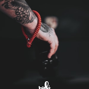 May include: A red camera strap made of braided cord with a metal clasp. The strap is worn on a tattooed arm.