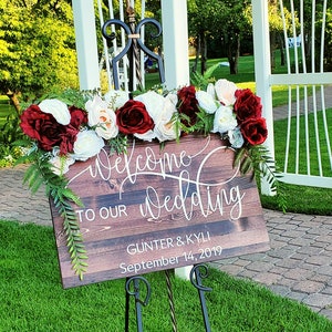 May include: A wooden sign with the text "Welcome to our Wedding" and the names "Gunter & Kyli" and the date "September 14, 2019". The sign is on an easel in front of a white wooden archway decorated with red and white flowers.