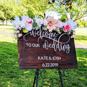 May include: A wooden wedding sign with white lettering that reads "Welcome to our wedding Kate & Josh 6.22.2019". The sign is decorated with pink and white flowers and greenery.