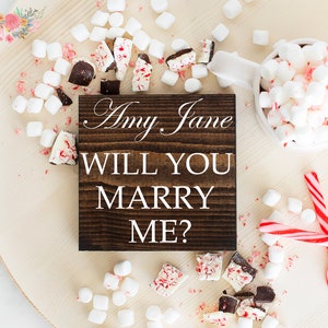 May include: A wooden sign with the text "Amy Jane Will You Marry Me?" surrounded by white marshmallows, candy canes, and chocolate pieces on a wooden surface.