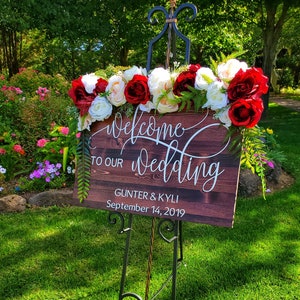 May include: Welcome to our wedding sign with red and white flowers on a wooden easel. The sign reads "Welcome to our wedding" with the names "Gunther & Kyli" and the date "September 14, 2019".