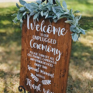 May include: A wooden sign with the text "Welcome to our Unplugged Ceremony" and a request to turn off all cell phones, cameras, and other devices. The sign is decorated with greenery and is displayed on a black easel.