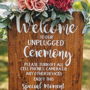 May include: A rustic wooden sign with the text "Welcome to our Unplugged Ceremony. Please turn off all cell phones, cameras & any other devices. Enjoy this special moment with us. Thank you." The sign is decorated with pink and red flowers and greenery.