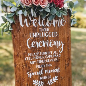 May include: A wooden sign with white text that reads "Welcome to our Unplugged Ceremony. Please turn off all cell phones, cameras & any other devices. Enjoy this special moment with us. Thank you". The sign is decorated with a floral arrangement of pink and red roses and greenery.