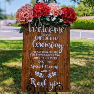 May include: A wooden sign with white text that reads "Welcome to our Unplugged Ceremony. Please turn off all cell phones, cameras & any other devices. Enjoy this special moment with us. Thank you". The sign is decorated with a floral arrangement of pink and red roses.