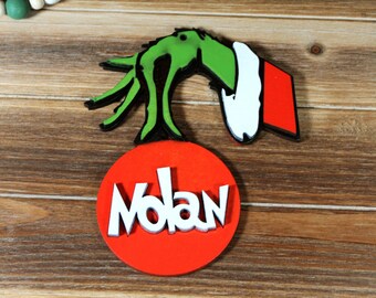 Puede incluir: Adorno de mano verde del Grinch con un gorro de Papá Noel rojo y blanco, y un círculo rojo con el nombre "Nolan" en letras blancas.