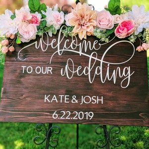 May include: A wooden wedding sign with a floral garland. The sign says "Welcome to our wedding" with the names "Kate & Josh" and the date "6.22.2019".