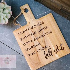 May include: A wooden cutting board with a handle and the text "Scary Movies, Pumpkin Spice, Cozy Sweaters, Murder, You Know... Fall Shit" engraved on it.