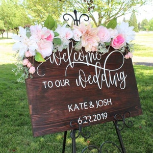 May include: A wooden wedding sign with white calligraphy that reads "Welcome to our wedding Kate & Josh 6.22.2019". The sign is decorated with pink and white flowers.