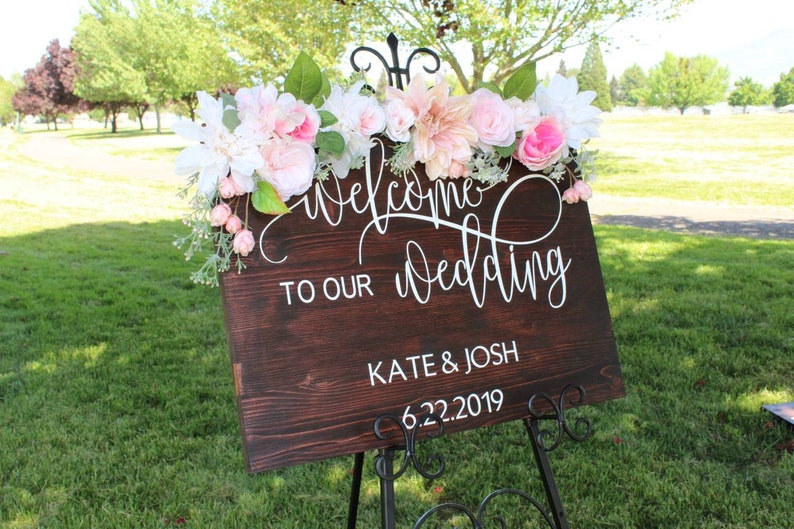 May include: A wooden wedding sign with white calligraphy that reads "Welcome to our wedding Kate & Josh 6.22.2019". The sign is decorated with pink and white flowers.