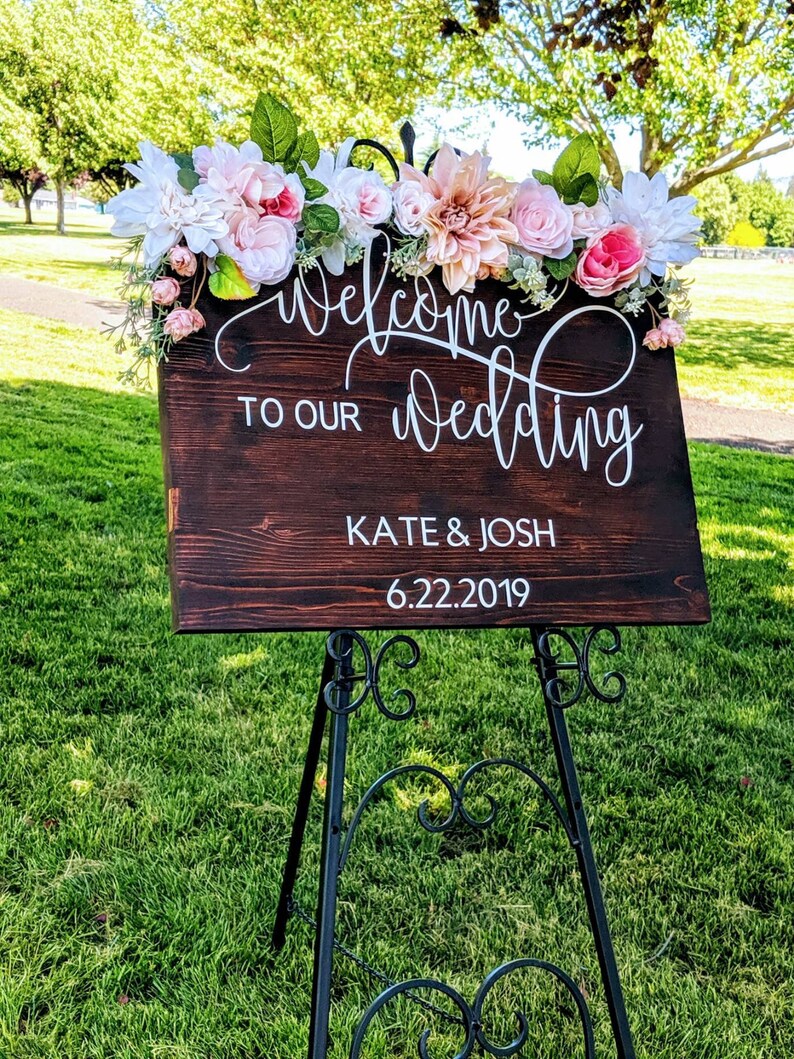 May include: A wooden wedding sign with white lettering that reads "Welcome to our wedding Kate & Josh 6.22.2019". The sign is decorated with pink and white flowers and greenery.