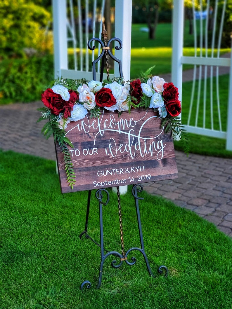 May include: Welcome to our wedding sign with red and white flowers on a wooden easel. The sign reads "Welcome to our wedding" with the names "Gunther & Kyli" and the date "September 14, 2019".