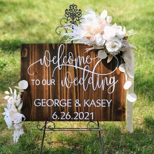 May include: A rustic wooden wedding welcome sign with white calligraphy that reads "Welcome to our wedding George & Kasey 6.26.2021". The sign is decorated with a floral arrangement of white and peach flowers and white feathers.