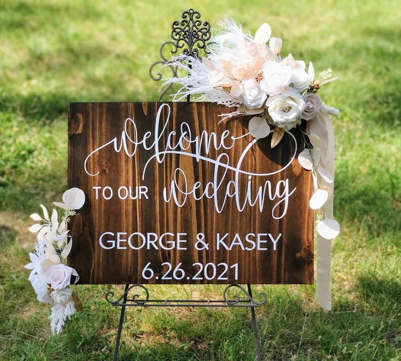 May include: A rustic wooden wedding welcome sign with white calligraphy that reads "Welcome to our wedding George & Kasey 6.26.2021". The sign is decorated with a floral arrangement of white and peach flowers and white feathers.