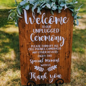 May include: A wooden sign with the text "Welcome to our Unplugged Ceremony. Please turn off all cell phones, cameras & any other devices. Enjoy this special moment with us. Thank you." The sign is decorated with a garland of green leaves.