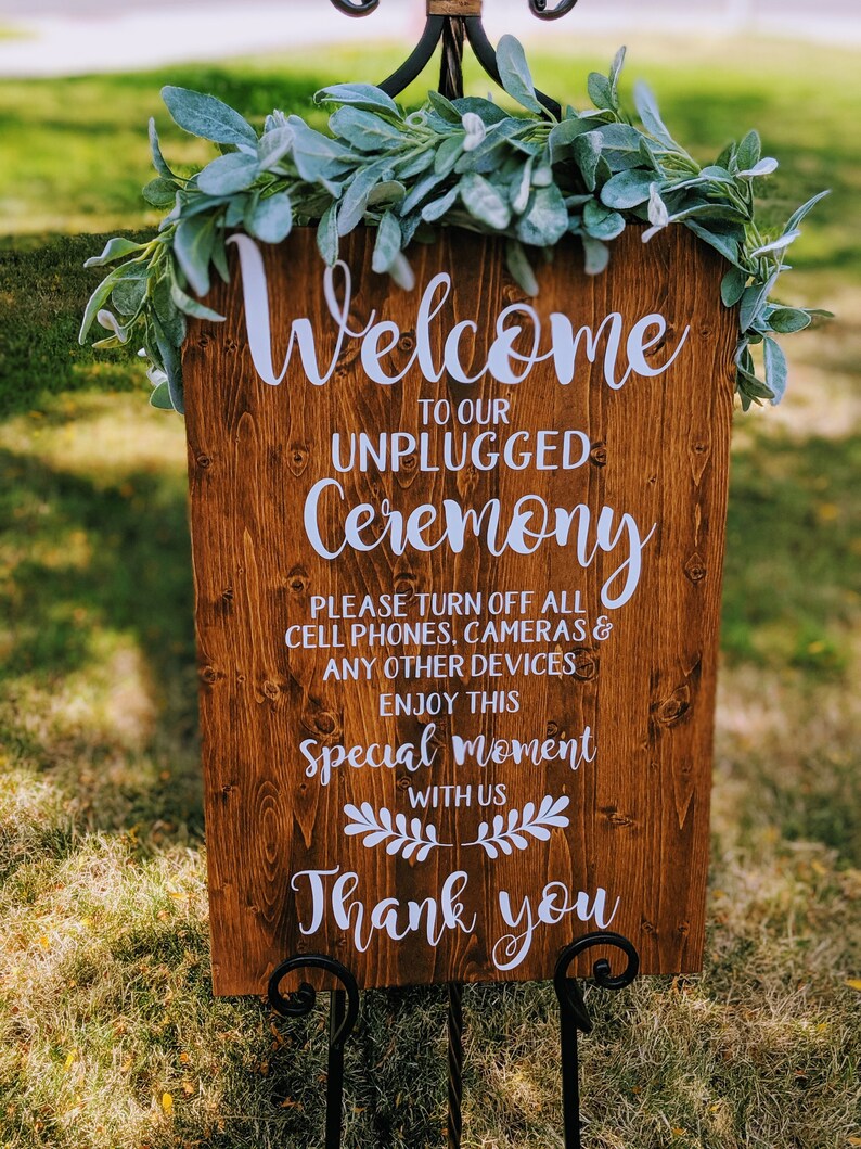 May include: A wooden sign with the text "Welcome to our Unplugged Ceremony. Please turn off all cell phones, cameras & any other devices. Enjoy this special moment with us. Thank you." The sign is decorated with a garland of green leaves.