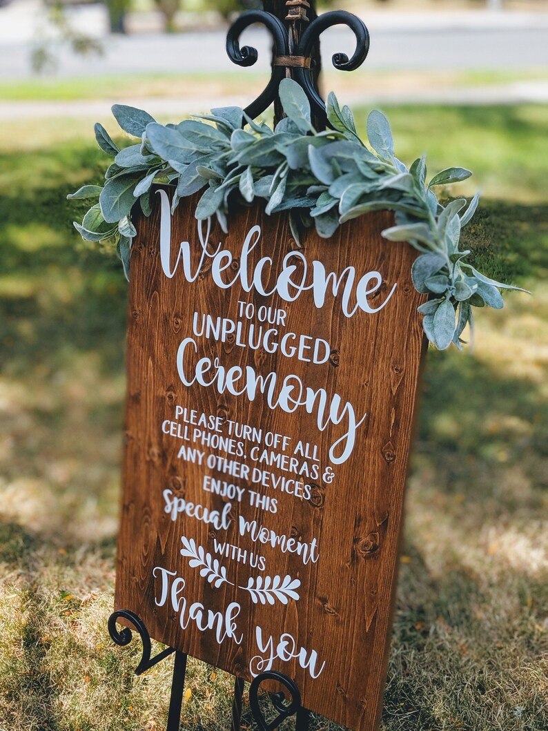 May include: A wooden sign with the text "Welcome to our Unplugged Ceremony" and a request to turn off all cell phones, cameras, and other devices. The sign is decorated with greenery and is displayed on a black easel.