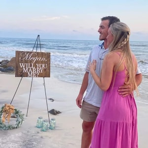 May include: A couple on a beach with a wooden sign that says "Megan, Will You Marry Me?" The sign is on a tripod and there are some glass jars in front of it.