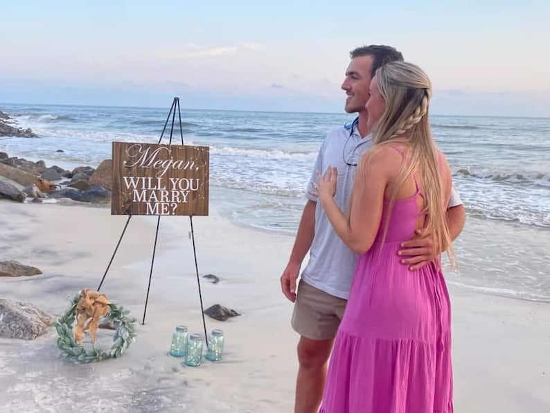 May include: A couple on a beach with a wooden sign that says "Megan, Will You Marry Me?" The sign is on a tripod and there are some glass jars in front of it.