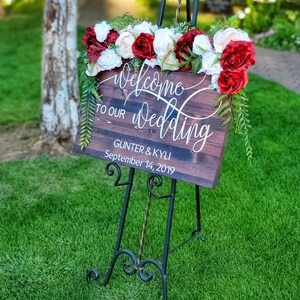 May include: Welcome to our wedding sign with red and white flowers on a wooden easel. The sign reads "Welcome to our wedding" with the names Gunter & Kyli and the date September 14, 2019.