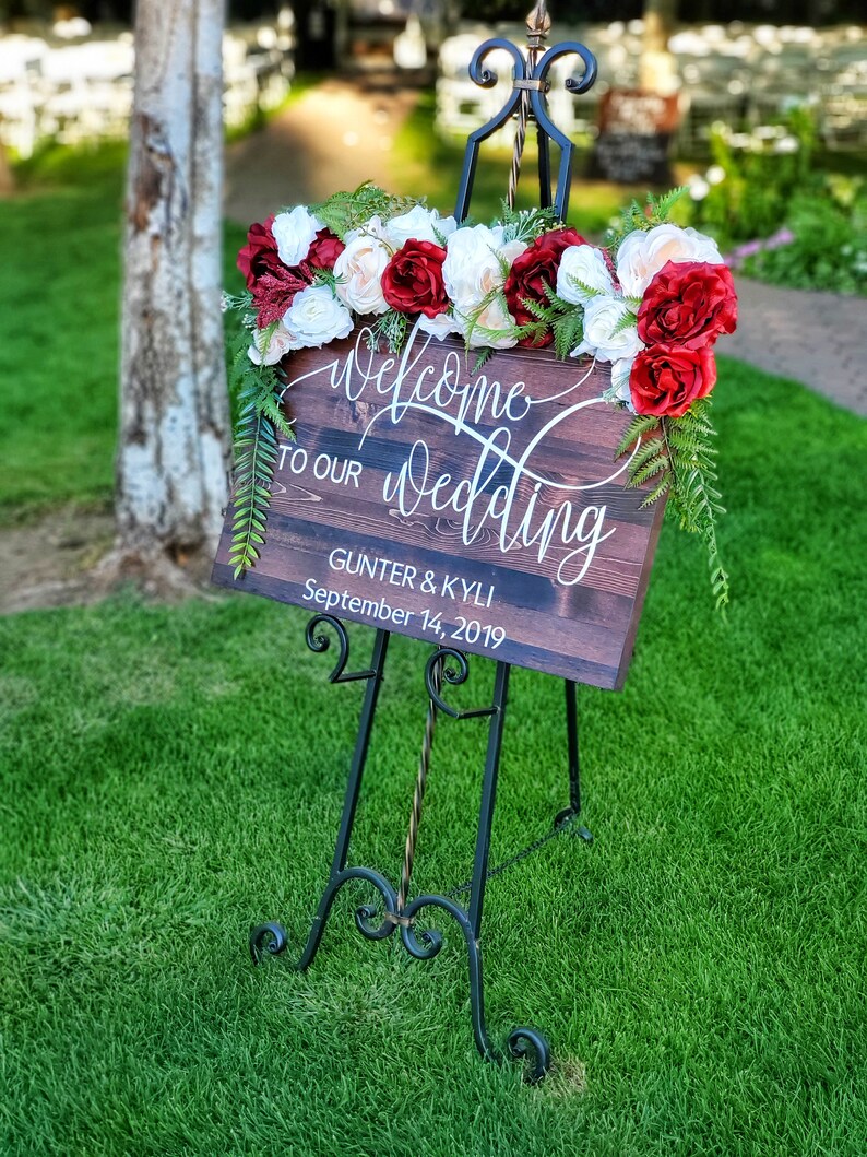 May include: Welcome to our wedding sign with red and white flowers on a wooden easel. The sign reads "Welcome to our wedding" with the names Gunter & Kyli and the date September 14, 2019.