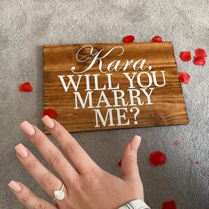 May include: A wooden sign with the words "Kara, Will You Marry Me?" in white lettering. A hand with a diamond ring is in the foreground.