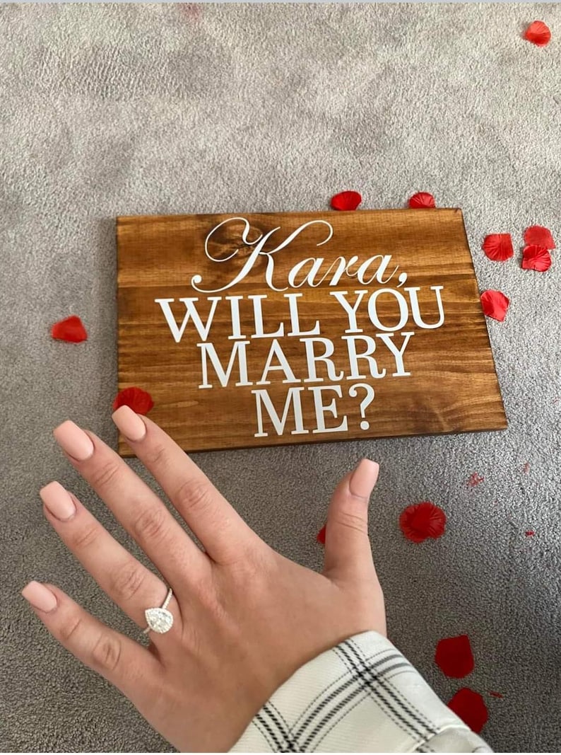 May include: A wooden sign with the words "Kara, Will You Marry Me?" in white lettering. A hand with a diamond ring is in the foreground.