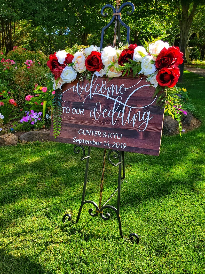 May include: Welcome to our wedding sign with red and white flowers on a wooden easel. The sign reads "Welcome to our wedding" with the names "Gunther & Kyli" and the date "September 14, 2019".