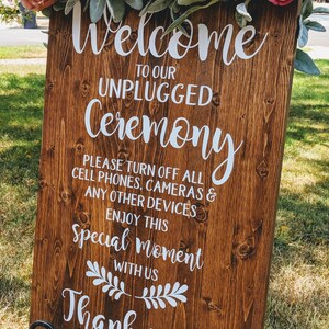 May include: A wooden sign with the text "Welcome to our Unplugged Ceremony. Please turn off all cell phones, cameras & any other devices. Enjoy this special moment with us. Thank you." The sign is decorated with a floral garland.